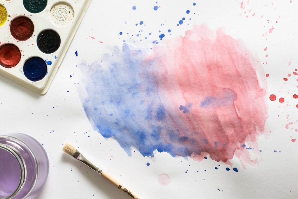 Benefits of Creative Arts in Early Childhood - A splattered painting of a blue and red heart shape is shown here with a brush and a pan of various colored watercolor paints.